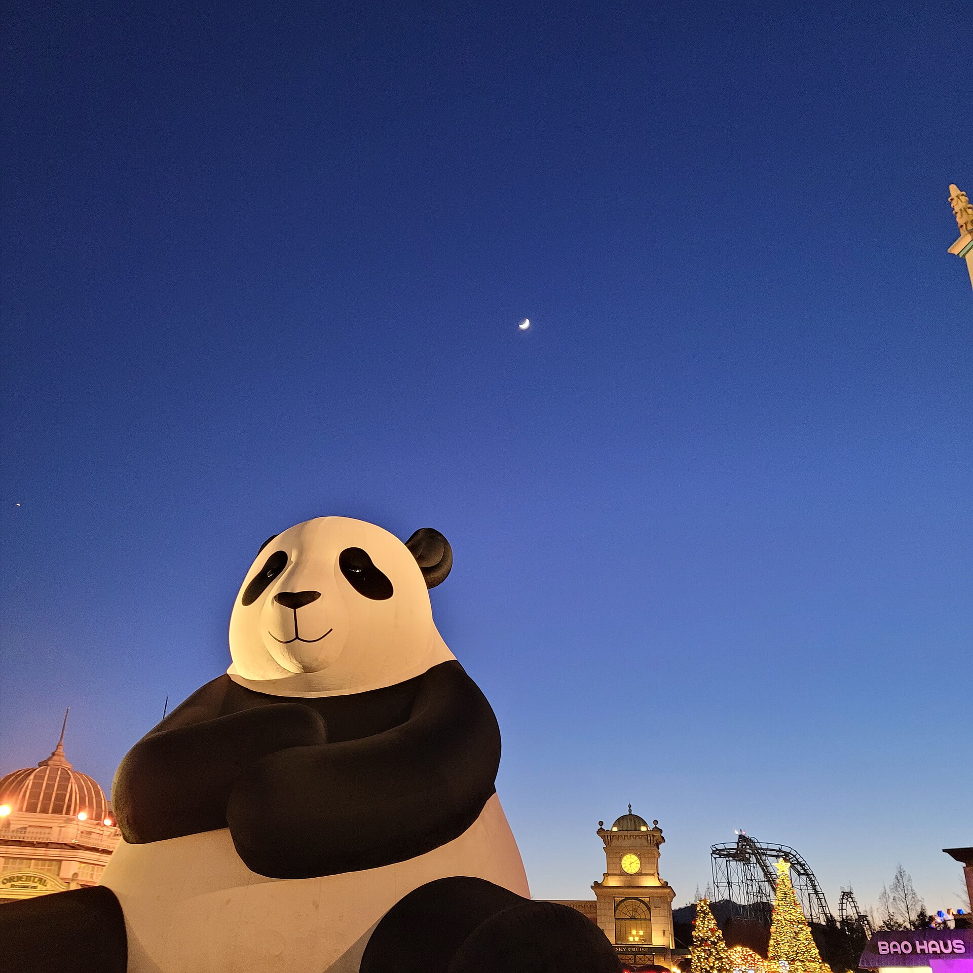 A giant panda statue with folded arms silhouetted against a deep blue evening sky with a crescent moon over Everland
