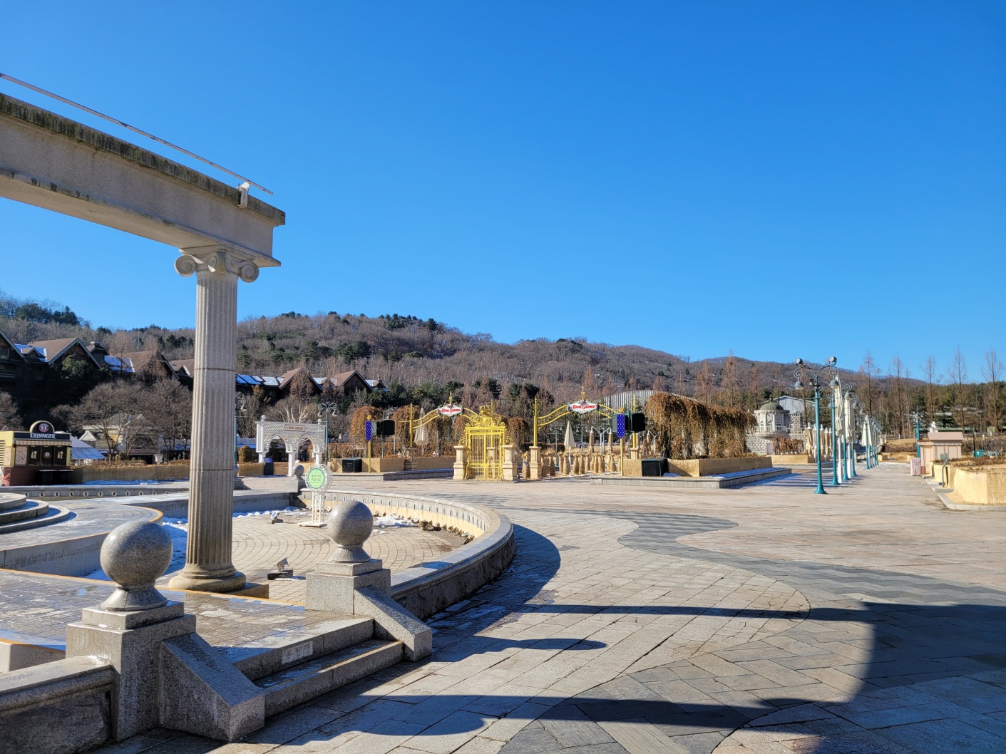 An empty terrace with classical columns and bare flower beds at Everland in winter, with leafless wooded hills behind under a blue sky