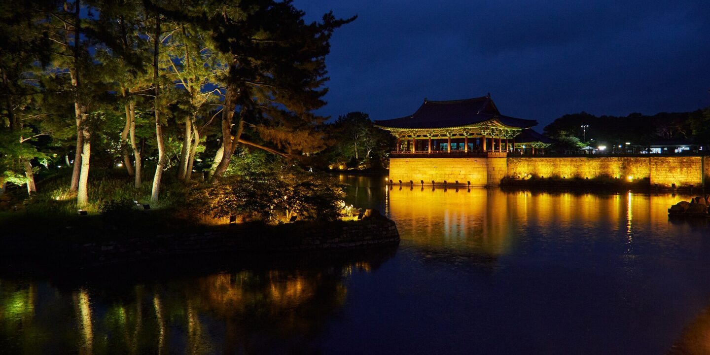 Floodlit pavilion of Donggung Palace reflected in Wolji Pond at dusk in Gyeongju, framed by pine trees