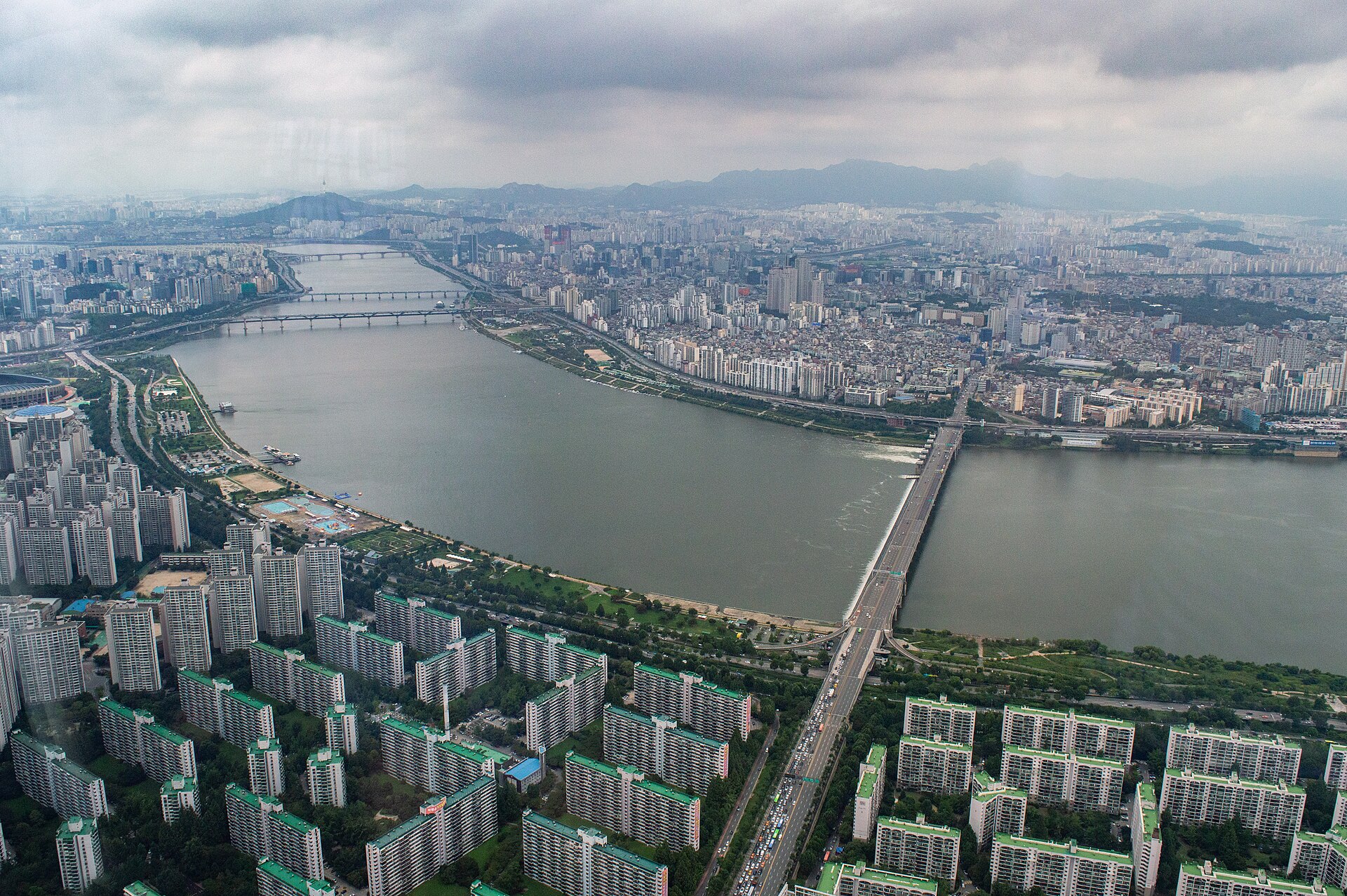 The Han River curving through Seoul seen from high above, with bridges, riverside parks and dense apartment blocks under a cloudy sky