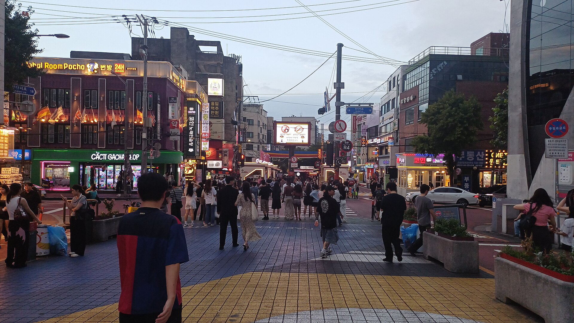 Busy Hongdae crossroads at dusk with illuminated shop signs and crowds of young people