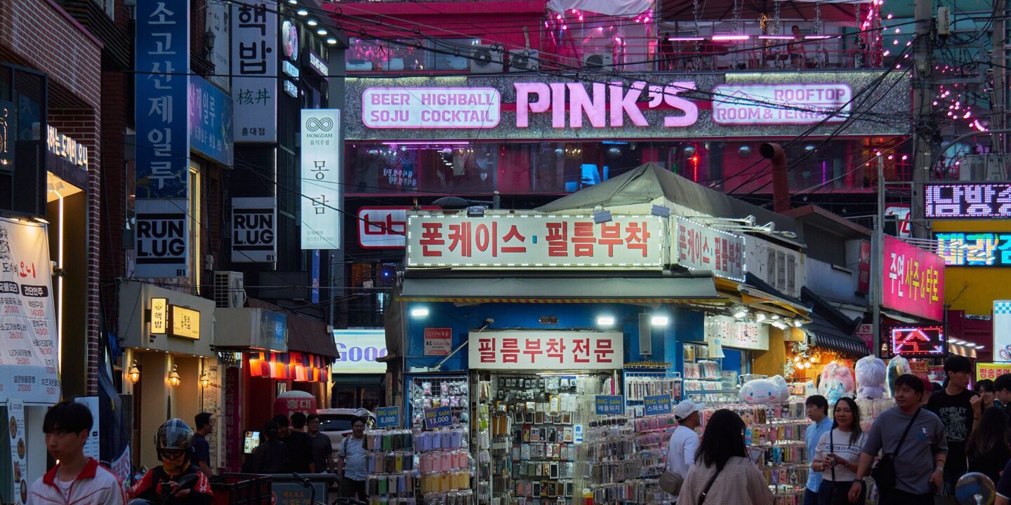 Hongdae street at dusk with neon signs, a rooftop bar and a phone accessory stall with shoppers