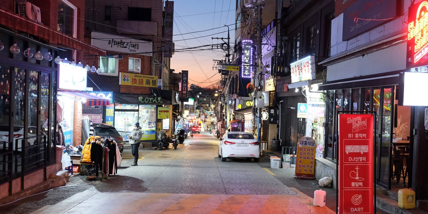 Itaewon side street at dusk with neon bar signs, a money exchange shop and a parked car