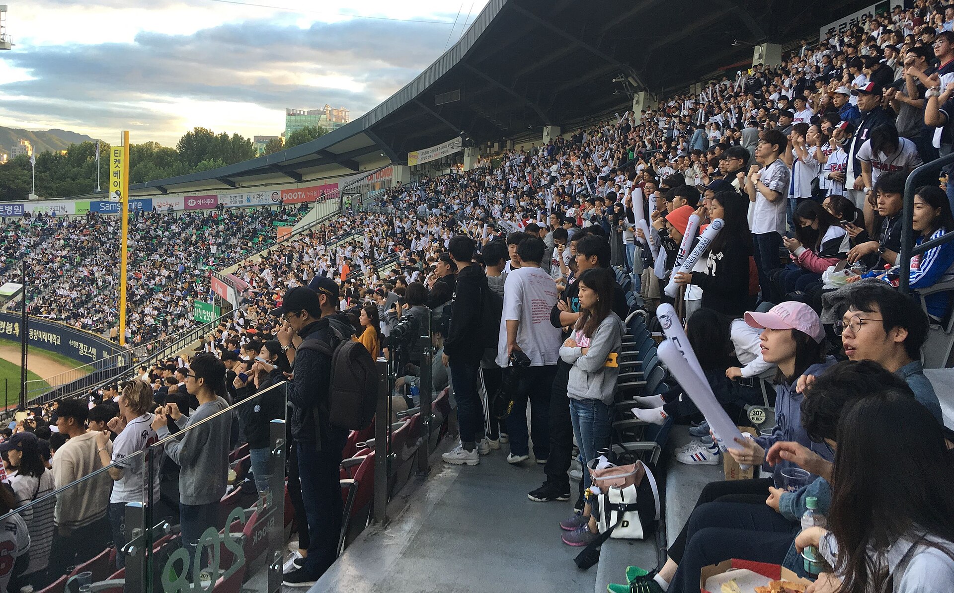 Packed stands of baseball supporters standing and waving inflatable thundersticks at Jamsil Stadium