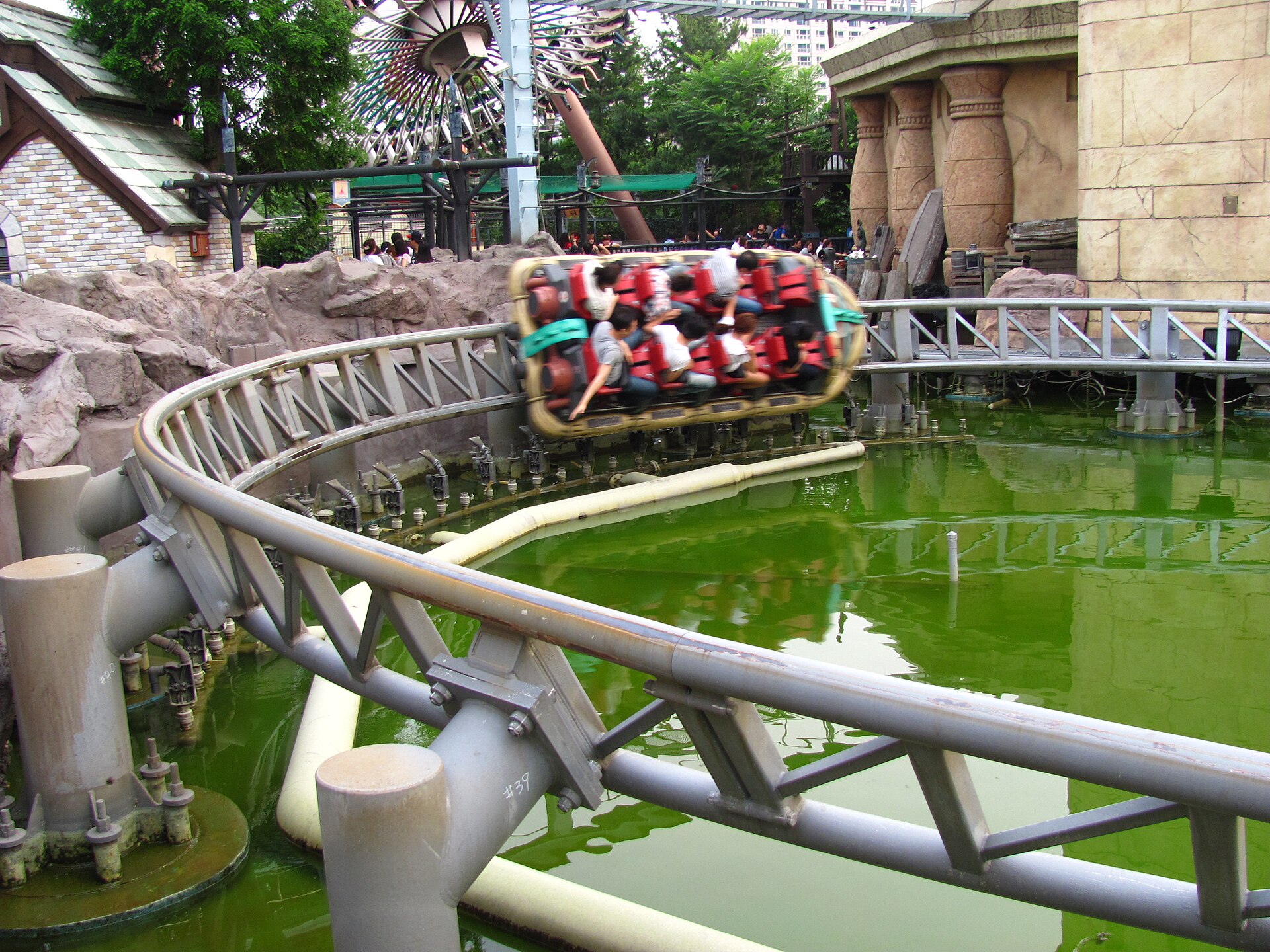 A carriage of riders on the Atlantis Adventure roller coaster racing along a track over green water beside themed ruins at Lotte World