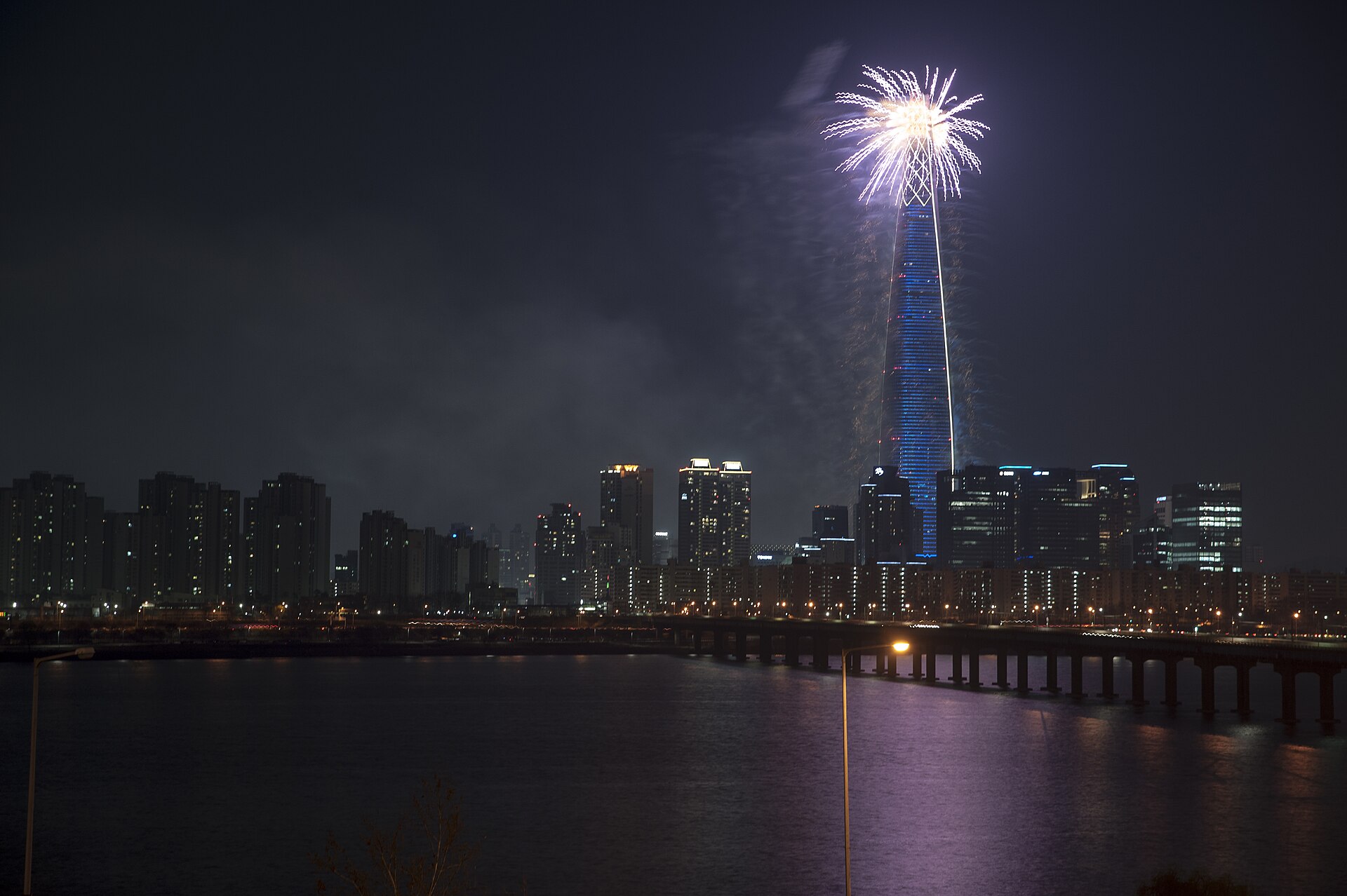 Fireworks bursting from the top of the illuminated Lotte World Tower at night, seen across the Han River with apartment blocks along the far bank
