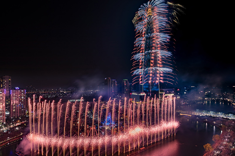 Fireworks cascading down Lotte World Tower above fountains on Seokchon Lake at night, with the city lit up around it