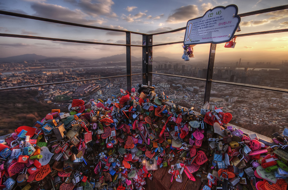 Thousands of colourful love padlocks piled on the terrace railings at N Seoul Tower, with the city spread out below at sunset