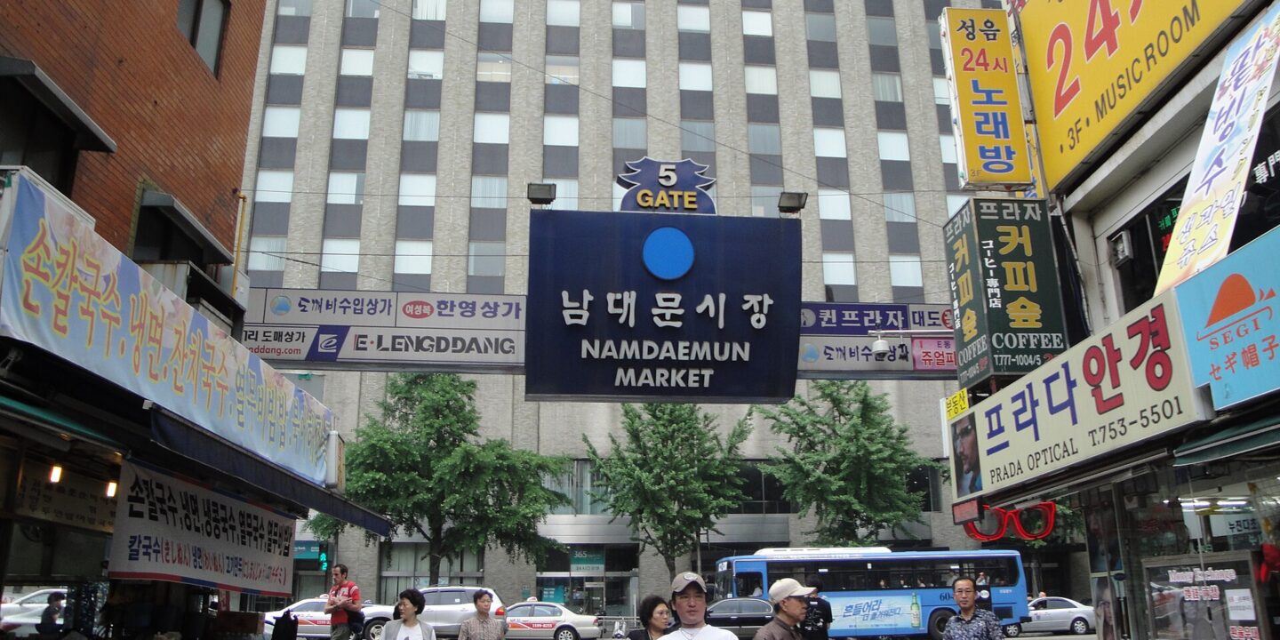 Namdaemun Market Gate 5 sign over a street with noodle stalls, optical shops and shoppers crossing the road