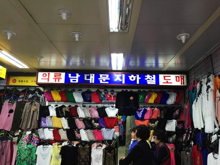 Underground clothing wholesale stall in Namdaemun with racks of colourful women's tops and a sign reading Namdaemun subway wholesale