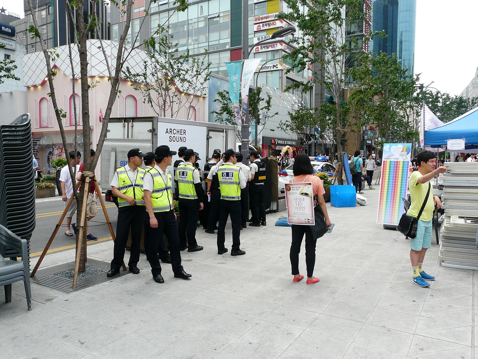 Police officers in high-visibility vests stand near festival stalls while a woman wears a Christian protest sign on a Seoul street