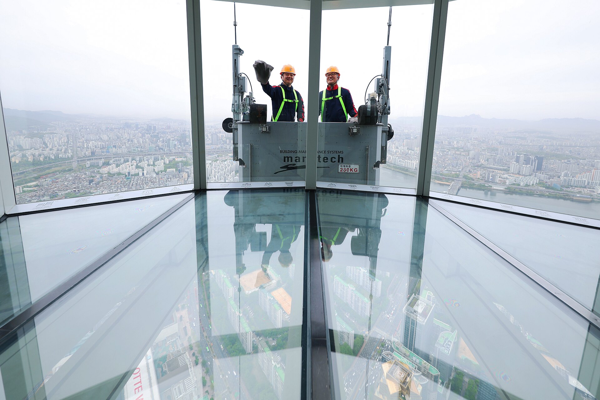 The glass floor of the Sky Deck at Seoul Sky, looking out through tall windows over the city, with two window cleaners in a cradle outside