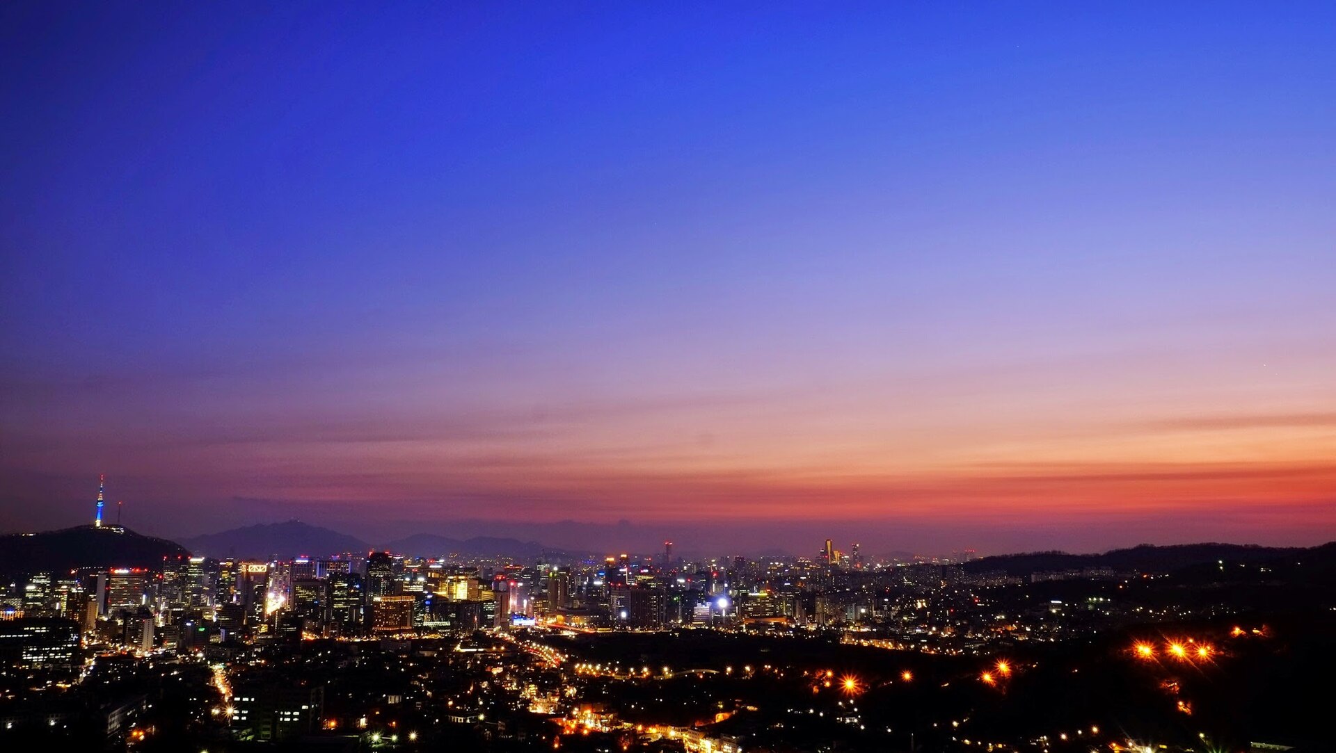 The Seoul skyline at dusk with N Seoul Tower silhouetted on its hill on the left under an orange and purple sky, city lights below