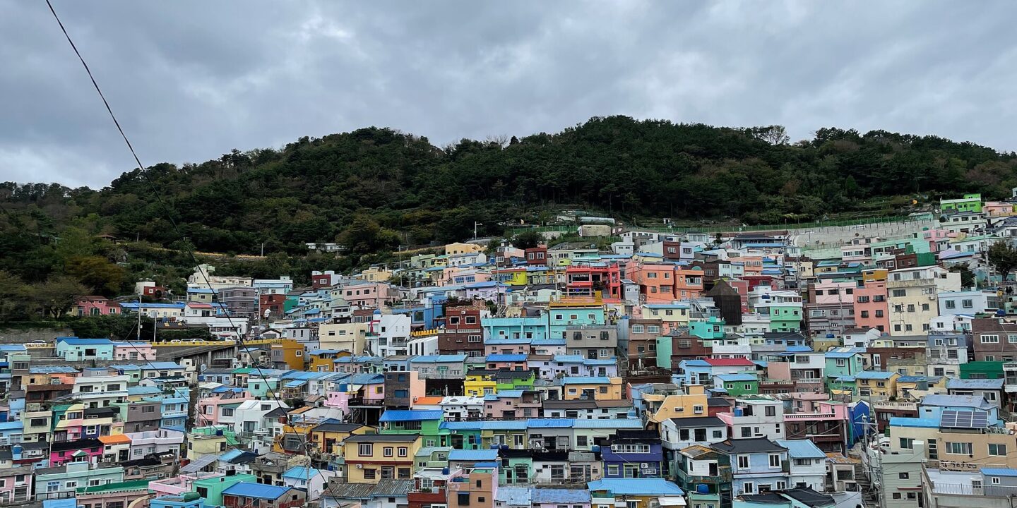 A hillside packed with small pastel-painted houses under a cloudy sky, with a forested ridge above
