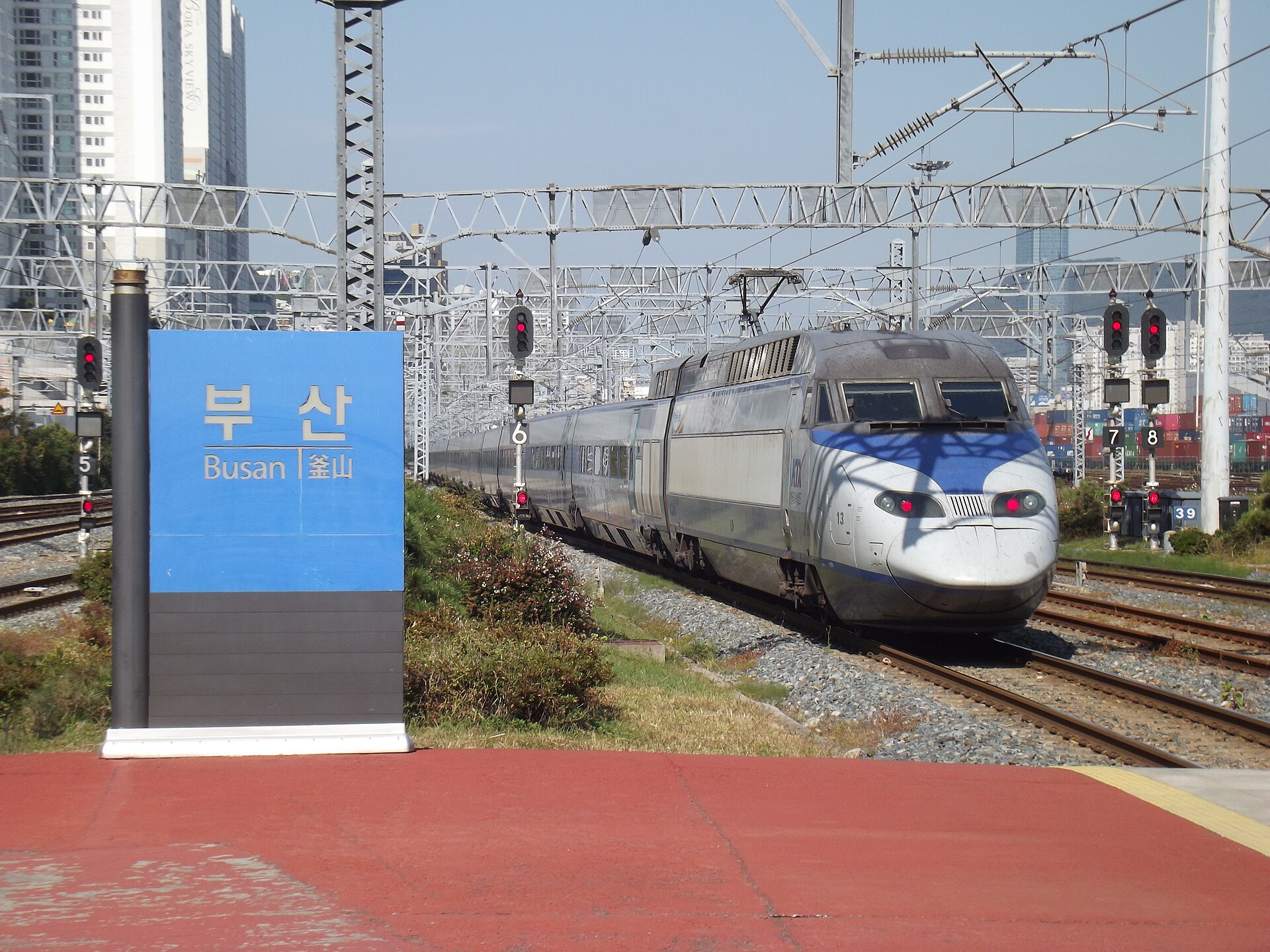 A blue station sign reading Busan in Korean and English on a platform, with a white high-speed train approaching on the tracks behind