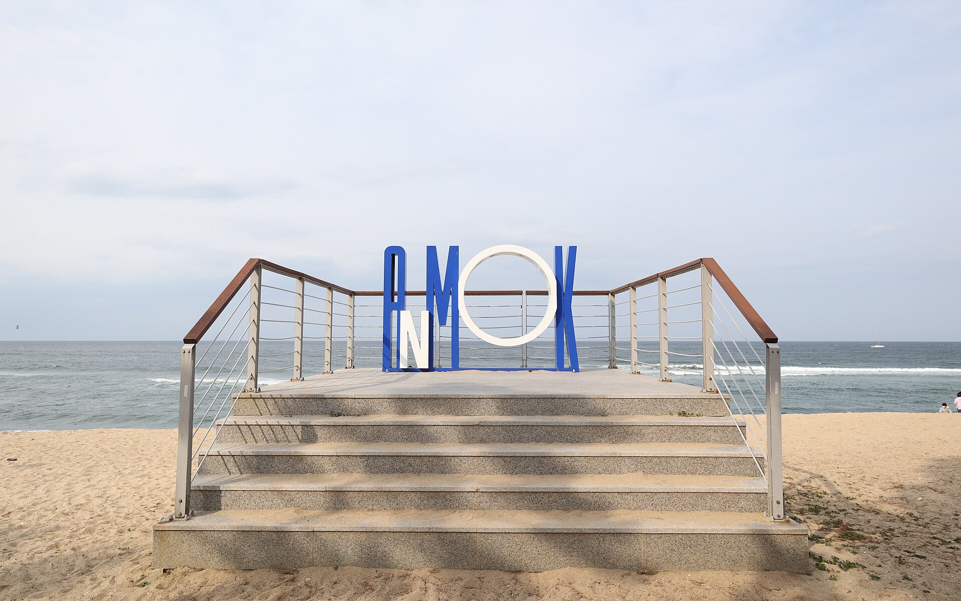 Large blue and white letters spelling ANMOK on a viewing platform with steps, on a sandy beach facing a grey sea