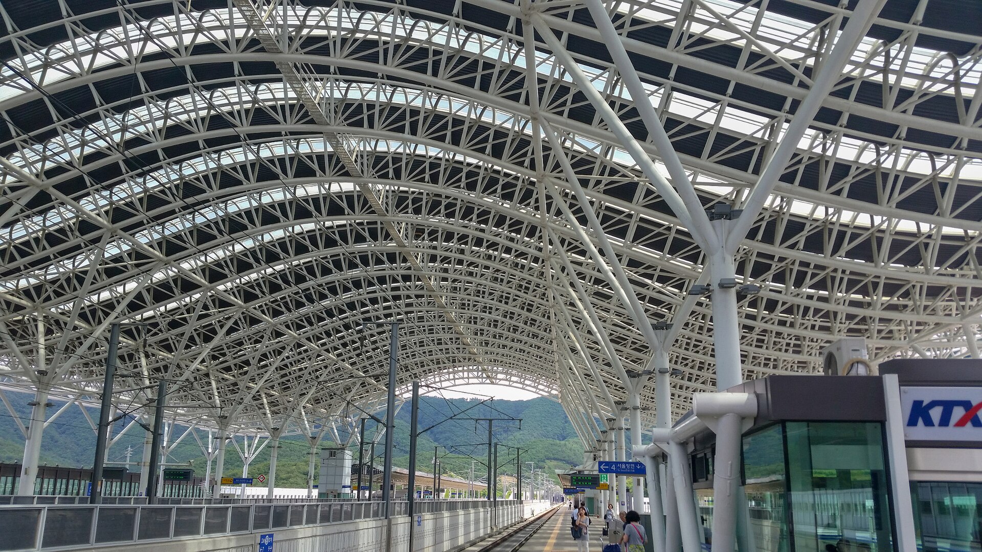 A railway station platform under a high white arched steel roof with glass panels, with a KTX sign on a building at the right