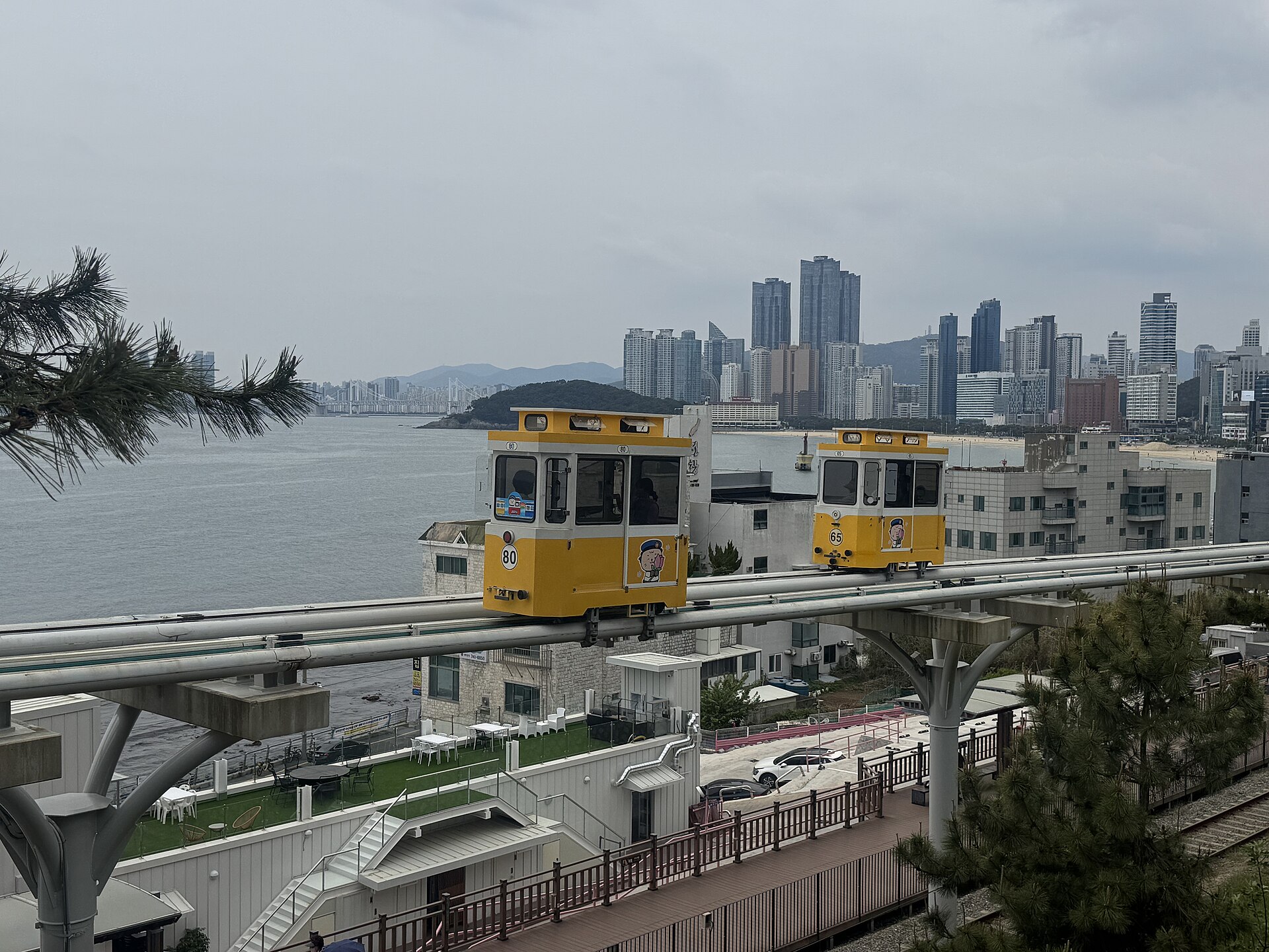 Two small yellow passenger pods on an elevated track above a coastal road, with the sea and a city skyline of tall towers behind