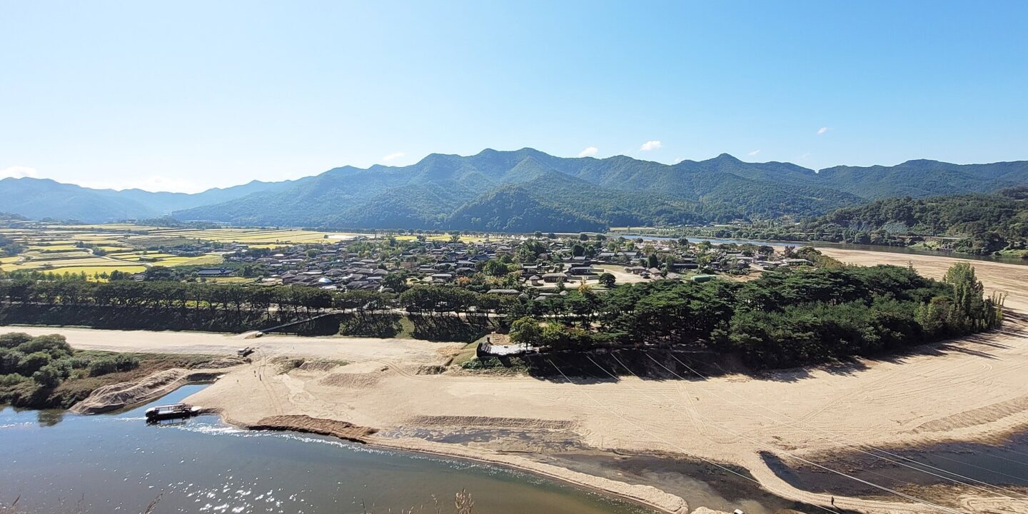 A traditional village of low roofs among trees and fields inside a wide bend of a shallow river, with mountains behind