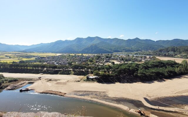 A traditional village of low roofs among trees and fields inside a wide bend of a shallow river, with mountains behind