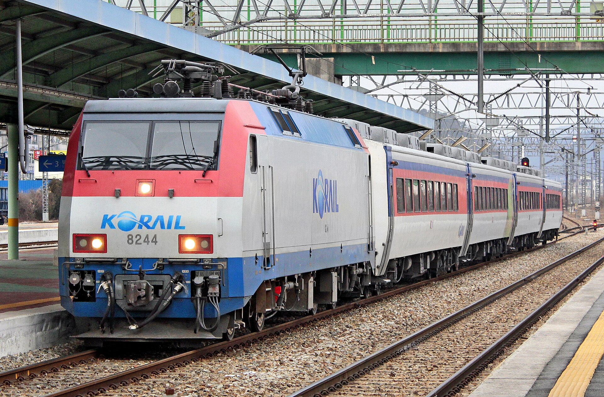 A red, white and blue electric locomotive with a KORAIL logo pulling passenger carriages past a station platform