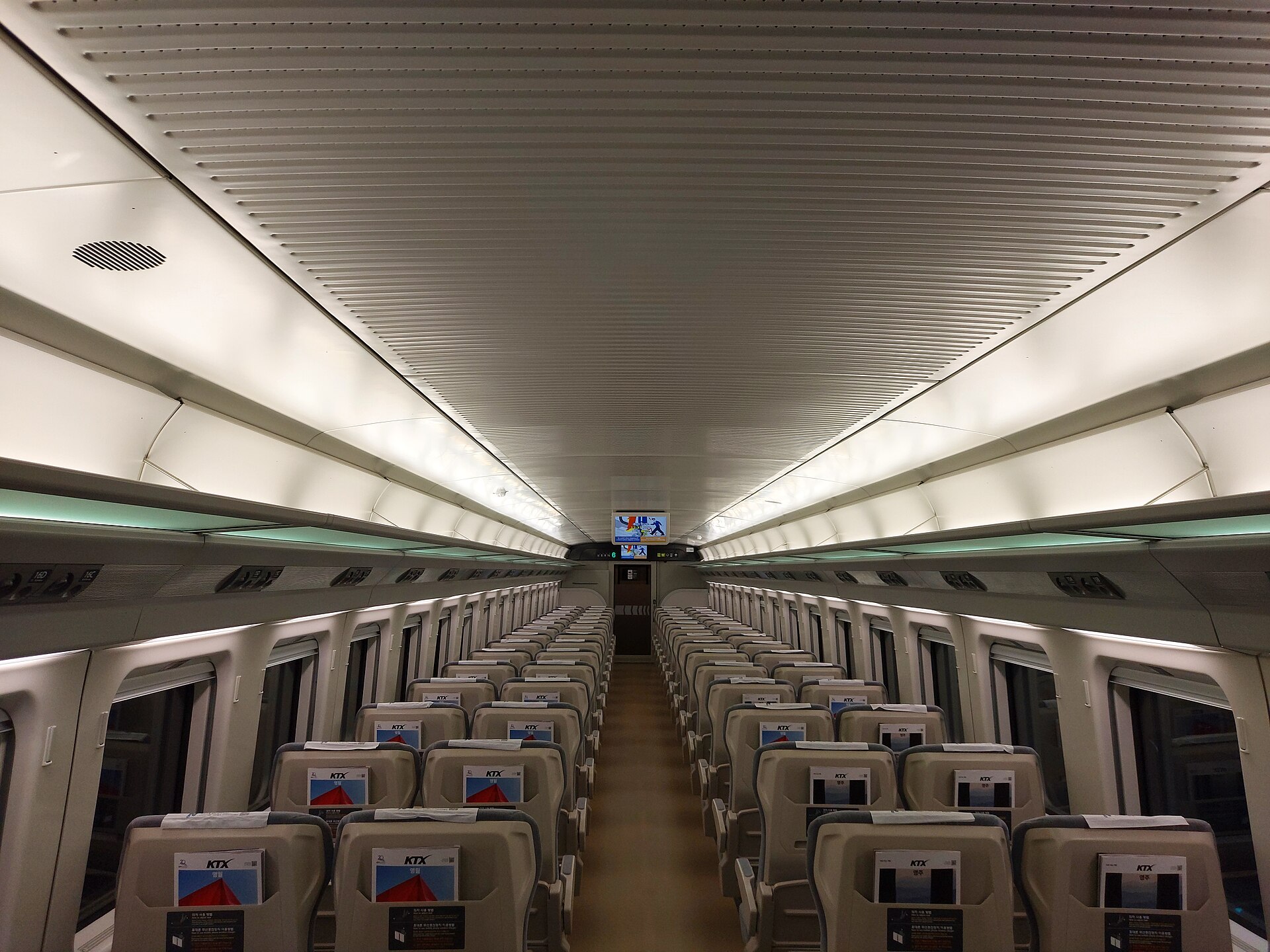 A long, bright high-speed train carriage with rows of cream seats with small screens in the seat backs and a central aisle