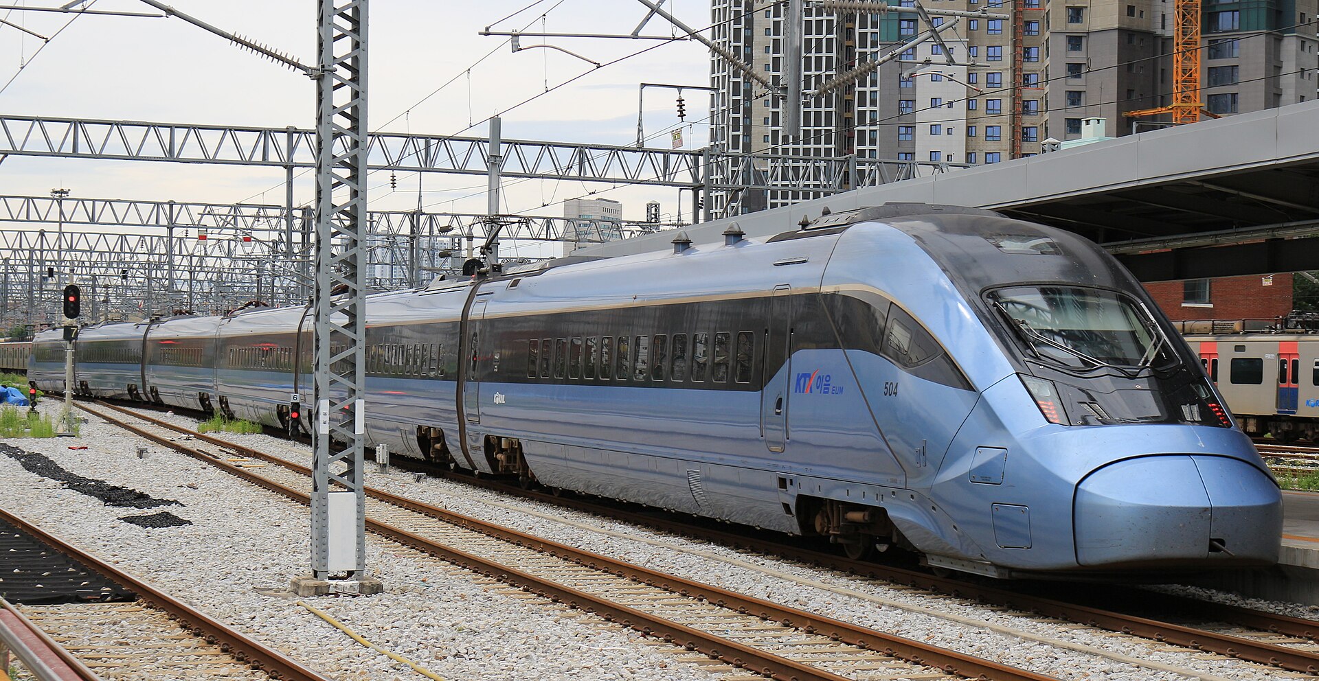 A pale blue high-speed train with a rounded nose at a station platform under overhead wires, with apartment towers behind