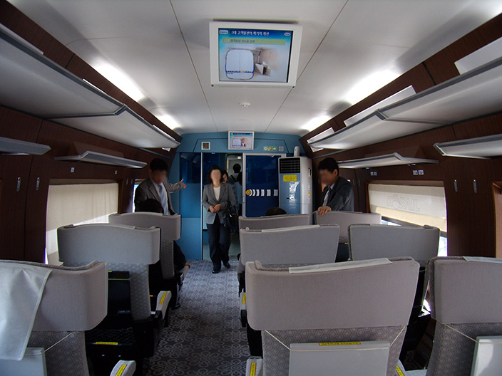 The interior of a first-class high-speed train carriage with wide grey seats in rows, a screen above the aisle and passengers walking down it