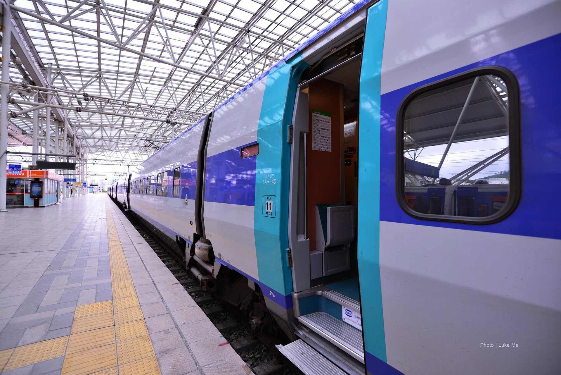 A white and blue high-speed train with an open door beside a long platform under a glass and steel station roof
