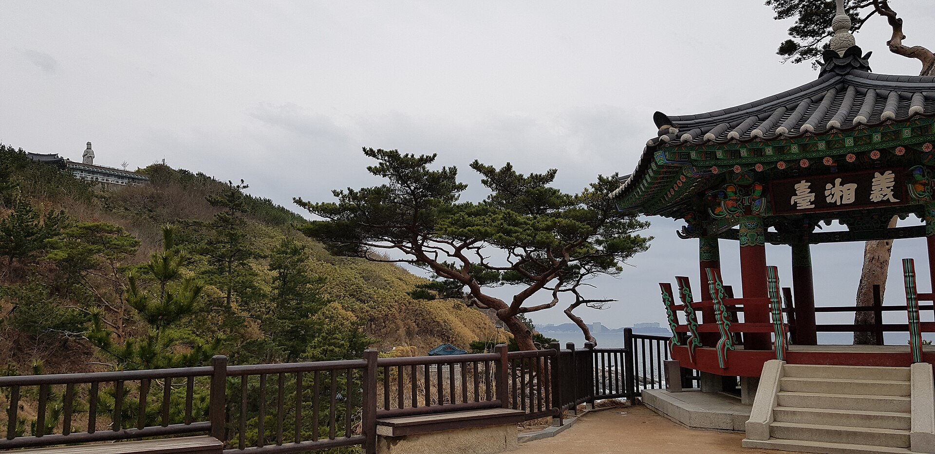 A small traditional pavilion with a painted wooden roof on a cliff edge beside twisted pine trees, with the sea visible beyond the railings