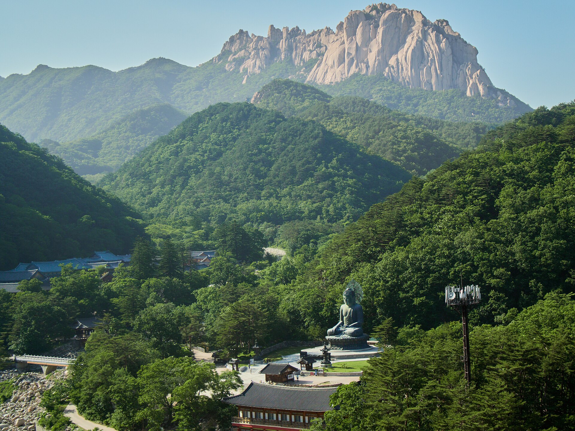 A large seated bronze Buddha in a temple courtyard surrounded by forested hills, with a long granite crag on the skyline behind