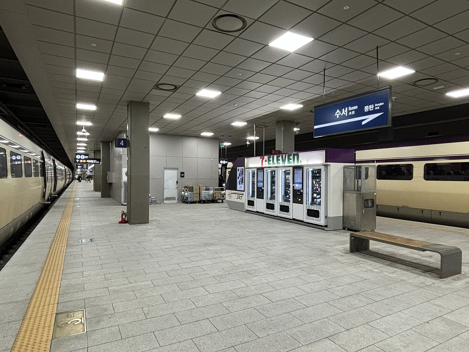 An underground railway platform with a 7-Eleven kiosk, a bench and a cream-coloured train on the left, under a station nameboard