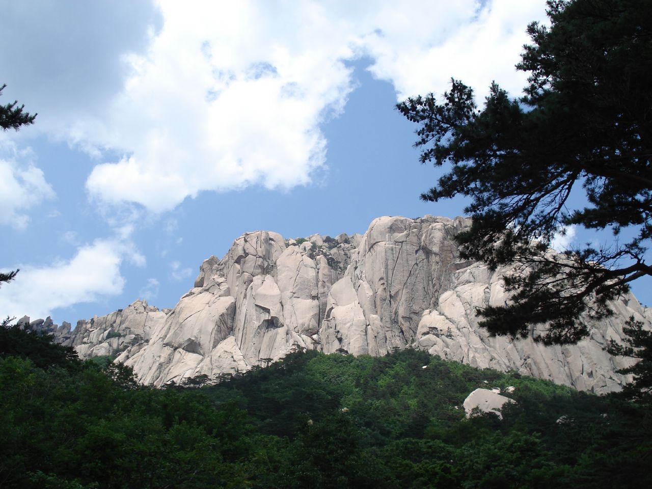 A long pale granite ridge with rounded and jagged sections rising above a dark green forested slope under a blue sky with white clouds