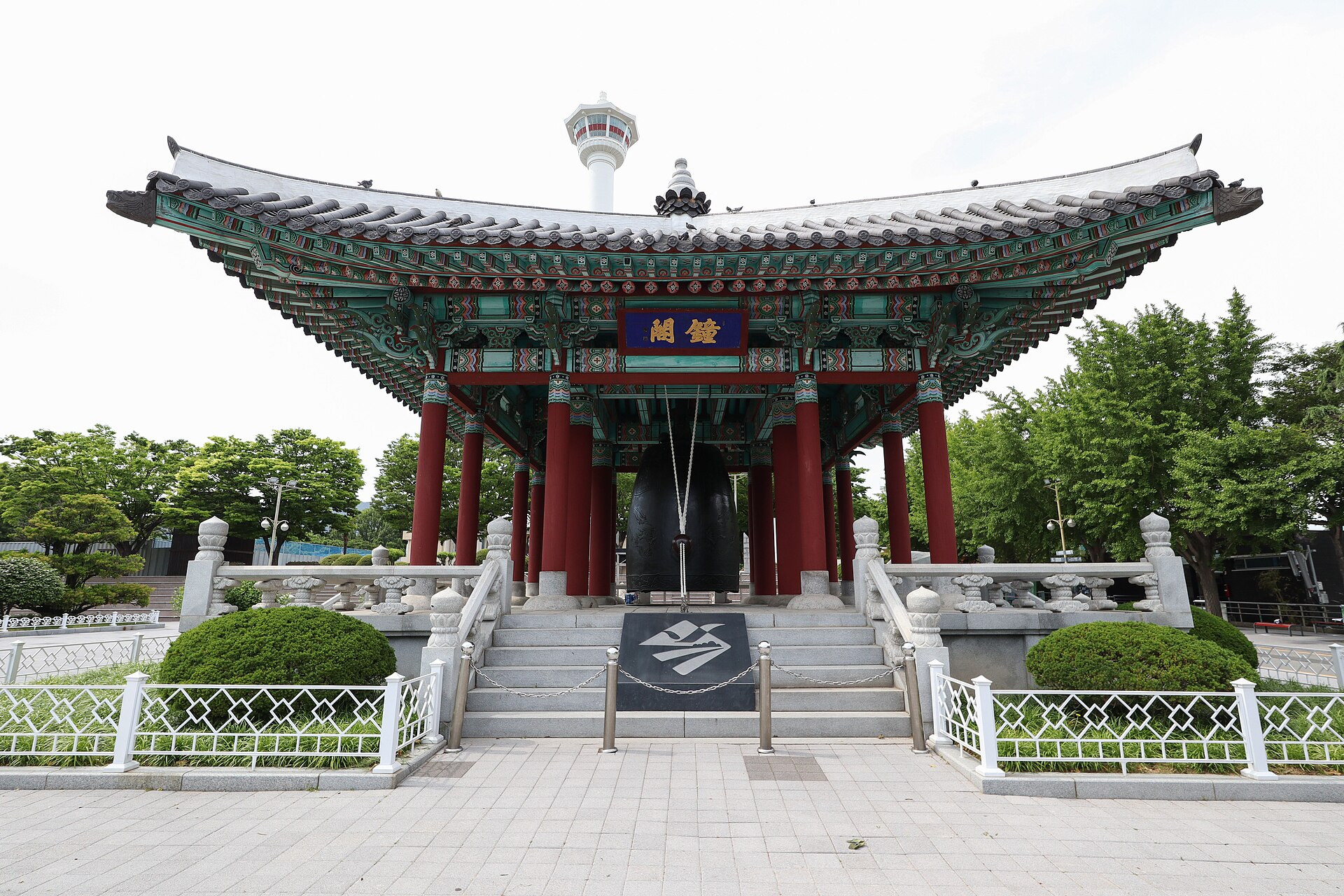 A traditional bell pavilion with a green and red painted roof in a paved park, with a white observation tower rising behind it